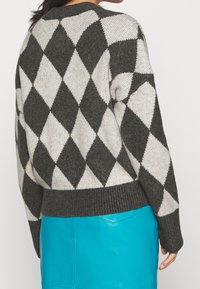 Maglione a maglia con motivo a diamante grigio e bianco, con orlo a costine, abbinato a una gonna lucida blu vivace. Il tessuto appare strutturato.