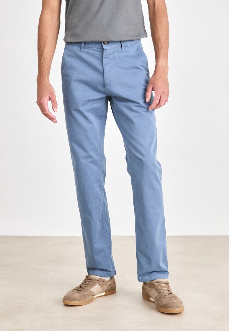 Homme portant un pantalon bleu clair et des baskets en daim beige, debout sur un sol clair avec un fond uni.