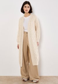 Langer beiger Strickcardigan mit strukturiertem Rautenmuster, offenem Frontbereich und gerippten Bündchen, getragen über einem weißen Oberteil und beigen weit geschnittenen Hosen.