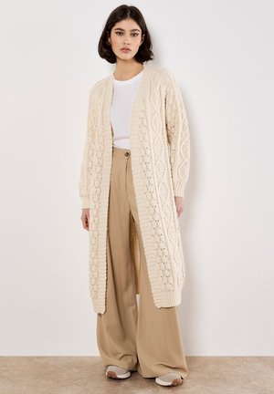 Langer beiger Strickcardigan mit strukturiertem Rautenmuster, offenem Frontbereich und gerippten Bündchen, getragen über einem weißen Oberteil und beigen weit geschnittenen Hosen.
