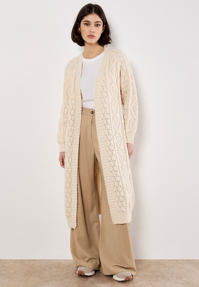 Langer beiger Strickcardigan mit strukturiertem Rautenmuster, offenem Frontbereich und gerippten Bündchen, getragen über einem weißen Oberteil und beigen weit geschnittenen Hosen.
