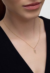 Złoty łańcuszek z delikatnym wisiorkiem w kształcie liścia, noszony na ciemnym materiale, podkreślającym jego skomplikowany wzór i blask.