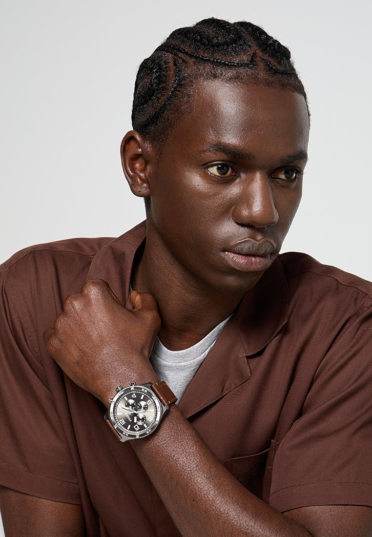 Brązowa koszula z krótkim rękawem, szary T-shirt, czarne warkocze. Zegarek na rękę z okrągłą, srebrną tarczą i brązowym skórzanym paskiem, z ciemnym cyferblatem i srebrnymi akcentami.