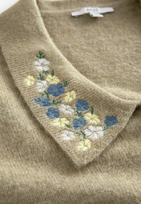 Stickad tröja med en beige textur och krage, med broderade blå, gula och vita blommor på vänster sida.