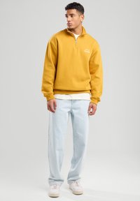 Yellow sweatshirt with a quarter-zip, featuring a crew neck and logo detail. Worn over a white shirt with light blue jeans and white sneakers.