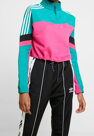 Osoba nosząca turkusowo-różową bluzę z suwakiem do ćwiartki i czarne spodnie dresowe Adidas z białymi paskami i logo.