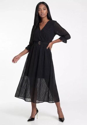 Kobieta w czarnej sukience midi z przezroczystymi wzorzystymi rękawami i spódnicą, z paskiem w talii, ubrana w czarne buty na wysokim obcasie, pozująca na białym tle.