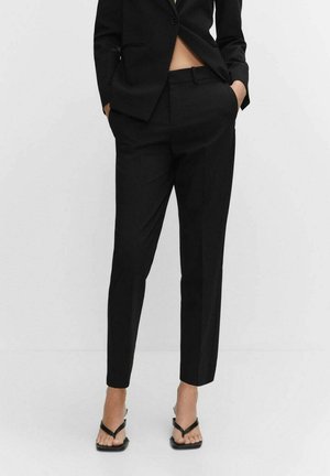 Femme portant un pantalon noir sur mesure et un blazer assorti, les mains dans les poches, associée à des sandales à talons noires à brides.