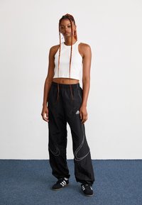 Sorte træningsbukser med hvidt svirl design og Adidas logo, parret med en tætsiddende hvid crop top. Modellen står på en blå tæppebaggrund.