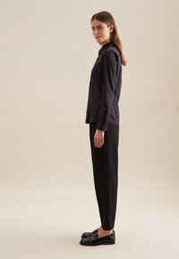 Camicia nera con bottoni, dotata di collo, abbinata a pantaloni neri. Il modello indossa mocassini neri lucidi. Tessuto liscio con dettagli minimi.