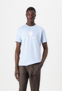 Ljusblått bomulls-t-shirt med rund hals och korta ärmar. Har en central vit "BELSTAFF"-logotyp och en grafisk emblem ovanför.