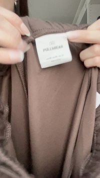 Material maro cu textură netedă, având o etichetă albă cu inscripția "PULL&BEAR." Eticheta conține instrucțiuni de întreținere și informații despre mărime.