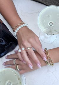 Två händer prydda med olika smycken: pärlarmband, guldringar, ett silverkedjearmband och ljusrosa manikyrerade naglar, på en marmor yta.