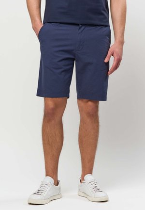 Man draagt marineblauwe shorts en witte sneakers, staat met één hand in de zak tegen een effen achtergrond.