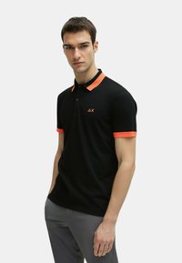 Polo nero con colletto e dettagli arancioni sulle maniche, dotato di un'apertura con tre bottoni e un piccolo logo sul petto. Tessuto dalla texture liscia.