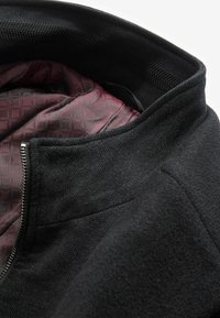 Schwarze Jacke mit strukturiertem Äußeren und glatter bordeauxfarbener Fütterung mit geometrischem Muster. Enthält einen hohen Kragen und einen Reißverschluss.
