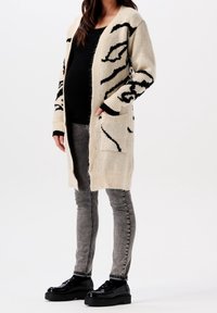 Pletený cardigan v béžové barvě s černým květinovým vzorem, se dlouhými rukávy, dvěma kapsami a volným střihem, doplněný o šedé skinny džíny a černé boty.