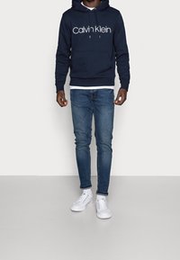 Man i marinblå Calvin Klein-hoodie, blå slim-fit jeans och vita Calvin Klein-sneakers mot en enkel ljus bakgrund.