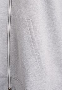 Sweat-shirt gris clair à fermeture éclair avec une texture douce, doté d'une poche latérale et d'une fermeture éclair lisse. Design minimaliste sans logos visibles.