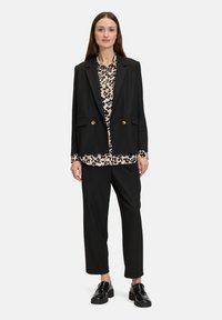 Schwarze taillierte Blazer mit goldenen Knöpfen über einer Bluse mit Leopardenmuster, kombiniert mit geraden schwarzen Hosen und schwarzen Slippern.