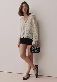Koronkowa bluzka z falbaniastym dołem, długimi bufkami, zestawiona z czarnymi szortami. Czarna torebka z kwiatowymi akcentami i ozdobnymi czarnymi balerinami.