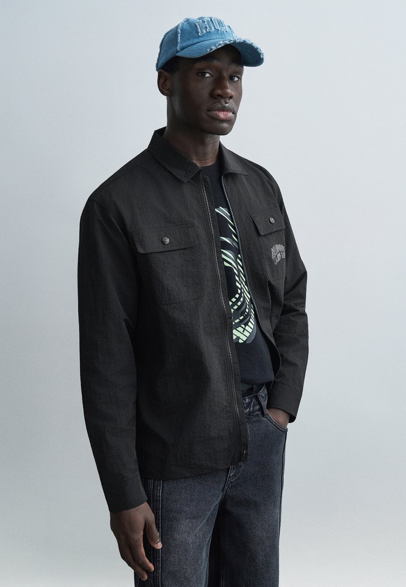 Μαύρο μπουφάν με φερμουάρ και δύο μπροστινές τσέπες πάνω από μαύρο μπλουζάκι με τύπωμα, σε συνδυασμό με σκούρα γκρι τζιν και ανοιχτό μπλε σκισμένο καπέλο.