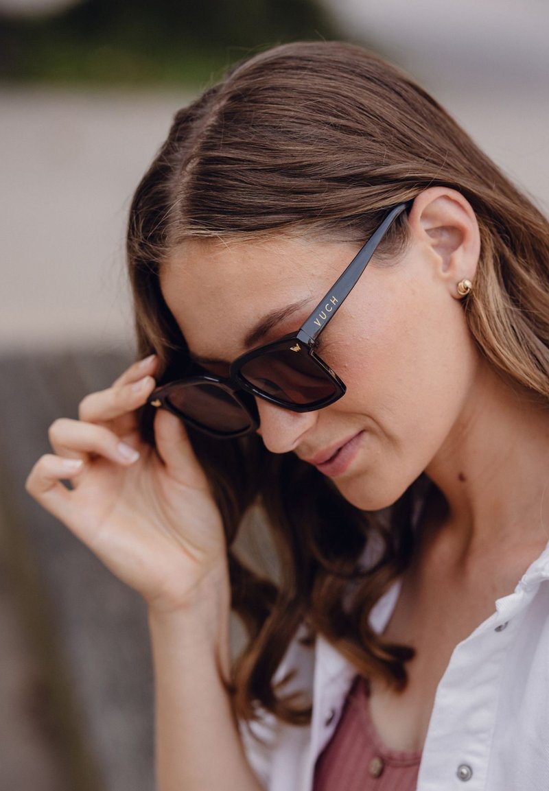 Černé obdélníkové sluneční brýle mají tmavé čočky a jemné zlaté logo na stranách. Rám má lesklý povrch.