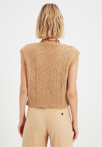Beiger Strickpullover mit kurzen Ärmeln, ausgestattet mit einem Zopfmuster und geripptem Saum. Tragen über beigen Hosen mit einer detailreichen Gesäßtasche.