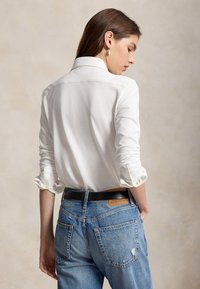 Mujer con cabello largo y castaño lleva una camisa blanca abotonada con las mangas remangadas y vaqueros azules de denim con un cinturón negro, de espaldas.