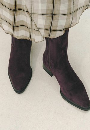 Purple suede ankle boots worn with a beige plaid skirt, shown against a pale neutral floor background.