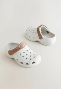 Sabots blancs à enfiler avec une sangle marron, dotés de trous de ventilation pour la respirabilité et d'une semelle structurée pour l'adhérence. Matériau en mousse.