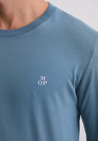 Niebieska koszulka z długim rękawem wykonana z gładkiej bawełny. Posiada okrągły dekolt oraz małe białe logo "MOP" na piersi. Prosty design, brak wzorów.