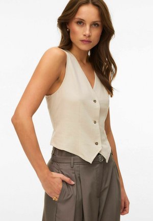 Kvinde iført en ærmeløs beige vest med knapper og højttaljede grå bukser med venstre hånd i lommen, foran en ensfarvet hvid baggrund.