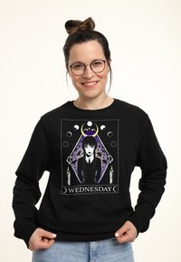 Felpa nera con una grafica di un personaggio con capelli lunghi davanti a un diamante viola, affiancato da candele e la scritta "WEDNESDAY."