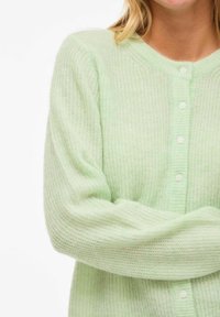 Gaiši zaļa adīta jakas ar mīkstu tekstūru, ar V veida kakla izgriezumu, garām piedurknēm un maziem baltiem pogām priekšpusē.