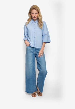 Jasnoniebieska koszula w paski na guziki z krótkimi rękawami, zestawiona z szerokimi dżinsami o luźnym kroju. Modelka stoi z rękami w kieszeniach, ma na sobie brązowe sandały.