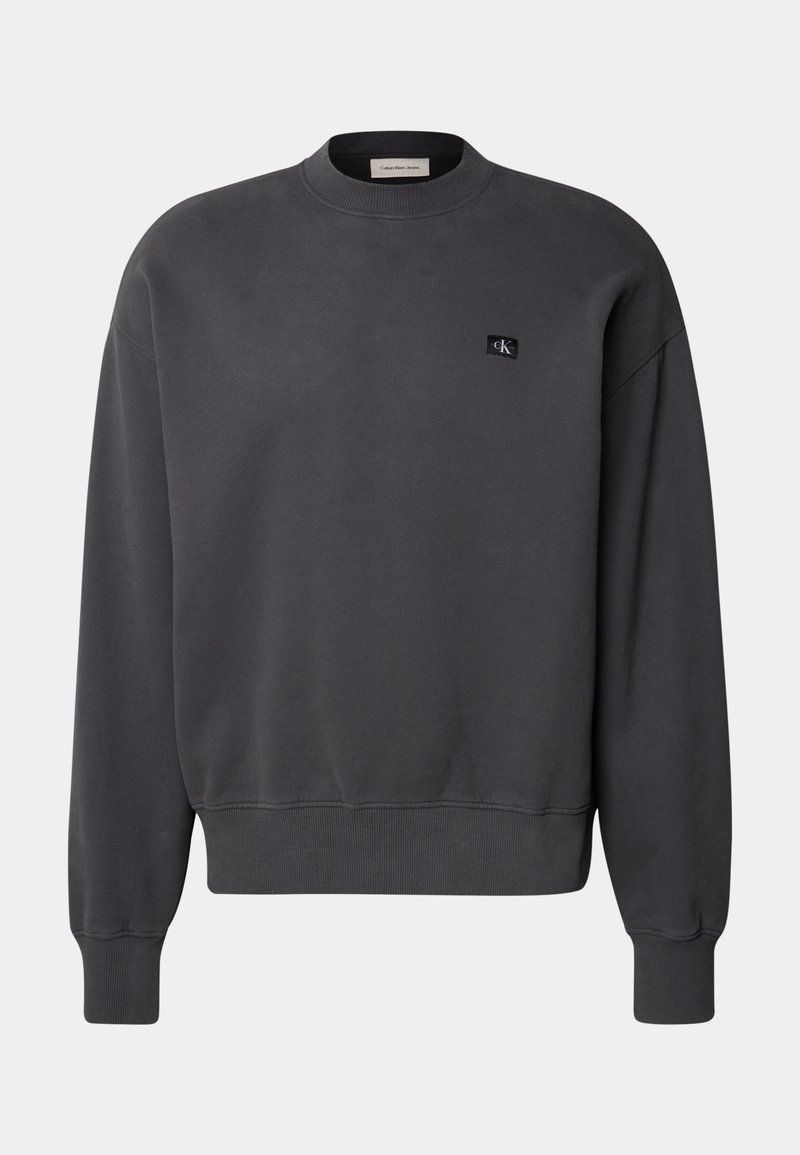 Sweat-shirt gris foncé en tissu doux, avec un col rond et des poignets côtelés, orné d'un petit patch logo noir sur la poitrine.