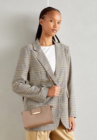 Blazer à carreaux en beige, noir et blanc ; coupe structurée avec revers. Tient un petit sac à main beige avec des finitions dorées. Fond clair.