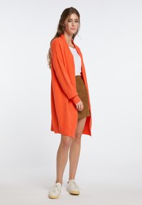 Orange lång kofta med öppen front, stickad textur och ribbade ärmslut, tillsammans med en vit t-shirt och en kjol i brunt mocka.