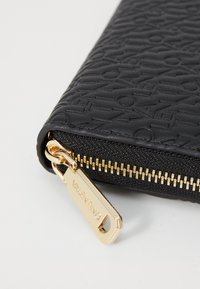 Black textured leather wallet featuring embossed branding, gold hardware zipper, and a rectangular shape with a prominent pull tab.