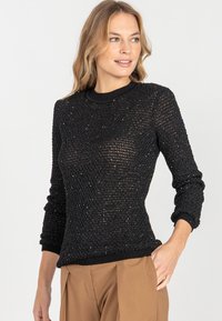 Kobieta ubrana w czarny sweter z długim rękawem, półprzezroczysty, dzianinowy, z delikatnymi błyskami, w połączeniu z brązowymi spodniami o wysokim stanie.