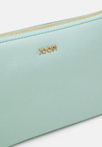 Pochette in pelle turchese chiaro con superficie testurizzata, zip dorata e logo: "JOOP!" impresso in oro sul fronte. Design pulito e semplice.
