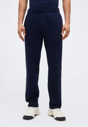 Homme portant un pantalon de sport bleu marine à bandes latérales et des baskets blanches, debout devant un fond clair uni.