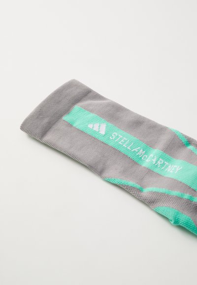 Calcetín deportivo gris con un patrón de malla texturizada. Presenta una banda de color teal claro con el logotipo "STELLA McCARTNEY" impreso en blanco.