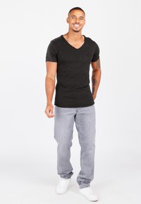 Zwarte V-hals T-shirt met korte mouwen, gecombineerd met lichtgrijze denim jeans en witte sneakers. Model staat voor een neutrale achtergrond.