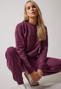 Sudadera púrpura y joggers a juego hechos de un tejido suave, con un corte relajado y puños acanalados. Combinados con zapatillas blancas.
