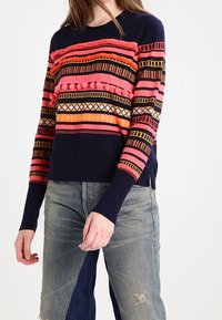 Femme portant un pull marine avec des rayures géométriques horizontales colorées et des pompons, associé à un jean large en patchwork.