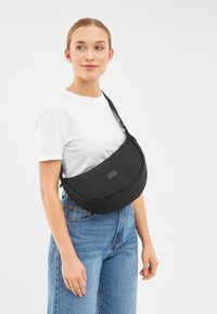 Borsa a spalla in nylon nero con forma curvata, tracolla regolabile e patch logo frontale. Presenta una texture liscia e un design minimalista.