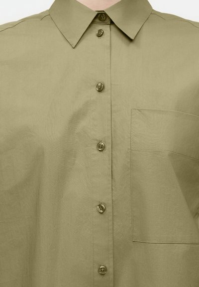 Πουκάμισο φερμουάρ σε ανοιχτό πράσινο ελιάς με μυτερό γιακά και μία τσέπη στο στήθος στην αριστερή πλευρά.