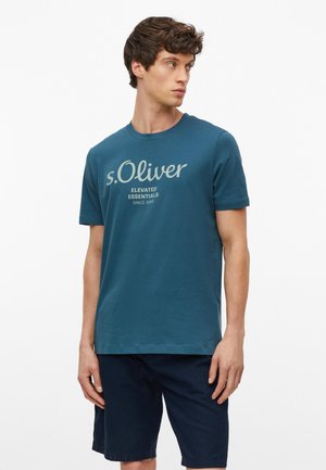 Młody mężczyzna w morskim t-shircie z napisem "s.Oliver" i granatowych szortach, stojący i patrzący na bok na jednolitym białym tle.